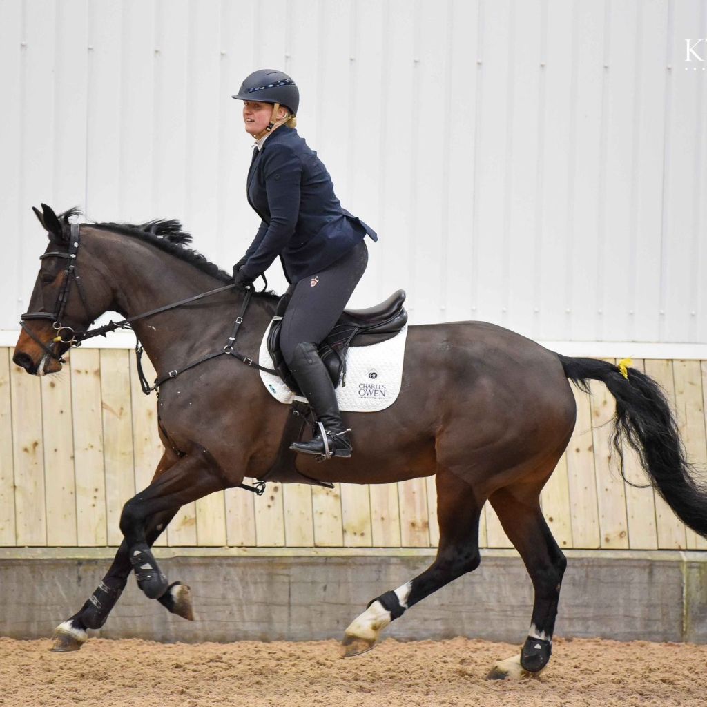 Laura Goodall riding a brown horse in an outdoor setting with a wooden fence in the background with her AV Edge horse bridle/collar patches (with EF technology, frequencies, not magnets, that may protect against modern technology EMFs and improve health and performance by stimulating the production of adenosine triphosphate/ATP)