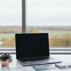 A silver laptop on a white desk in front of a window with a countryside view, featuring a yellow AV Edge EMF Protect Filter applied to the palm rest. This EF Technology solution is designed for work-from-home wellness, potentially helping to reduce tech-fatigue and brain fog while maintaining a high-performance digital workspace.
