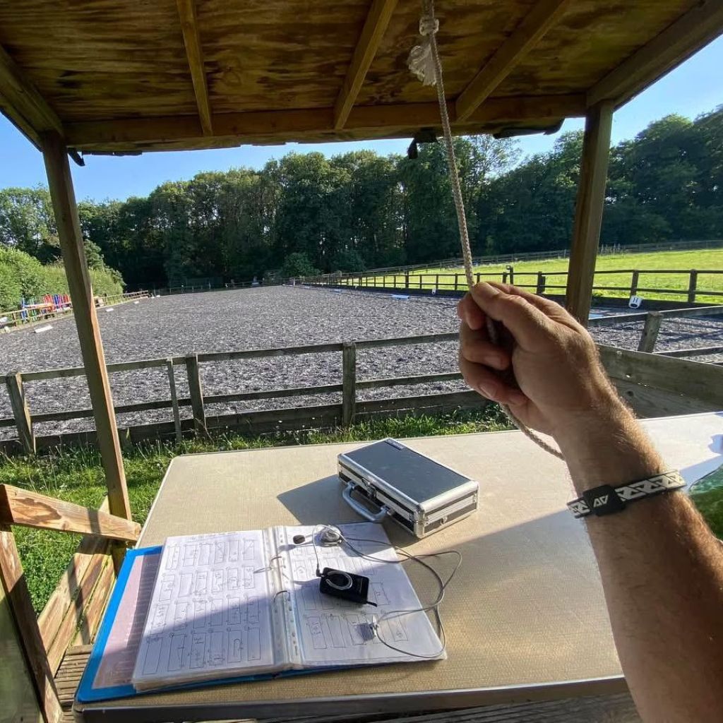 Alex Gingell dressage judge holding a string over a table with a notebook, solar panel, and camera on a wooden platform overlooking a field wearing his AV Edge Legend Wristband with with EF technology, frequencies, not magnets, that may protect against modern technology EMFs and improve health and performance by stimulating the production of adenosine triphosphate/ATP