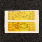 A close-up shot of two rectangular, golden Cell Phone EMF Protect Filters on their adhesive backing. The holograms feature a geometric star and circle pattern, highlighting the EF Technology used as a frequency-based filter to provide biological EMF protection for mobile devices.