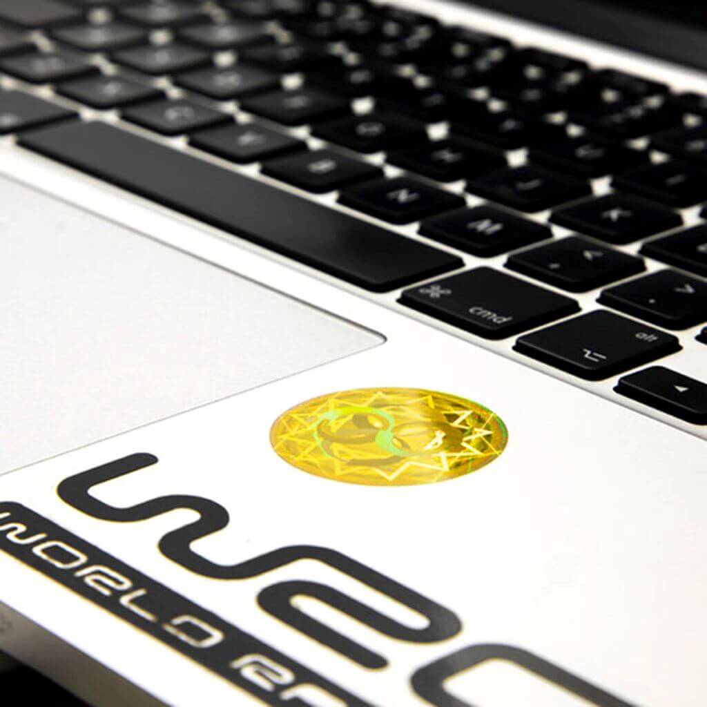A close-up, angled shot of a silver laptop keyboard and palm rest, featuring a circular, yellow AV Edge EMF Protect Filter applied near the trackpad. The frequency-based EF Technology is designed to provide a biological filter for modern technology frequencies, potentially helping to reduce brain fog and tech-fatigue for the modern professional.