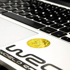 A close-up, angled shot of a silver laptop keyboard and palm rest, featuring a circular, yellow AV Edge EMF Protect Filter applied near the trackpad. The frequency-based EF Technology is designed to provide a biological filter for modern technology frequencies, potentially helping to reduce brain fog and tech-fatigue for the modern professional.